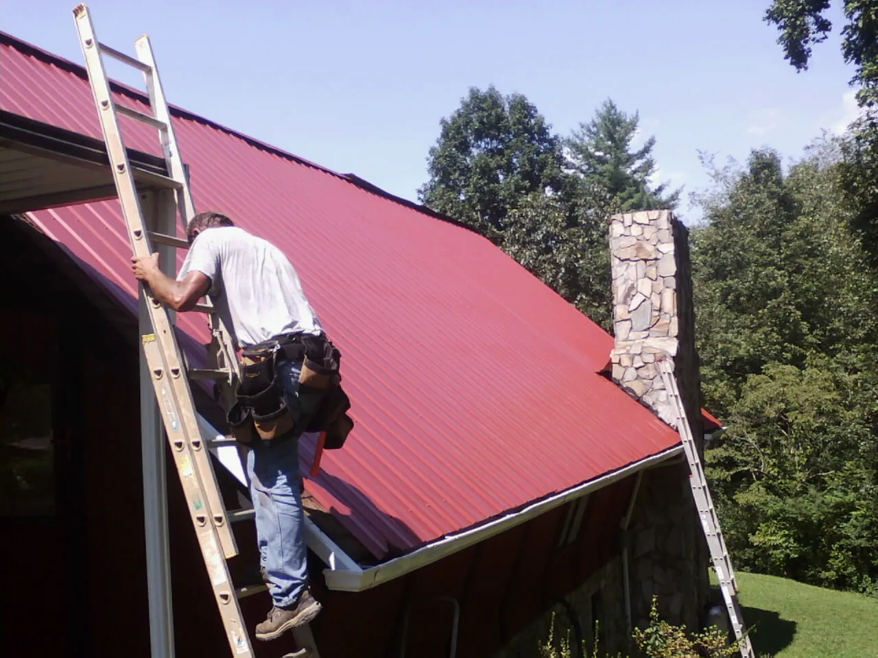 Roofer on a ladder beside a red metal roof and stone chimney — Soffit & Fascia Repair by Roofing Brothers in Woods Hole
